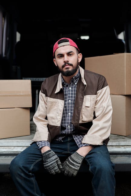 A young male mover with a beard and wearing a black cap and a white polo shirt stands inside the open rear of a moving van, with arms crossed and a serious expression. The interior of the van is fille