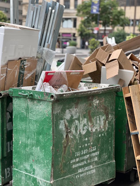 A large, green industrial waste disposal bin filled with various household and packaging materials, including cardboard boxes, plastic waste, and some metal items. The bin is placed outdoors on a paved area, with a mixture of opened and crushed cardboard boxes spilling over the top. In the background, there are additional cardboard boxes, some wooden pallets, and a street scene with trees and buildings, indicating a busy urban environment. The image captures a home relocation or moving process involving loading or disposal of bulky waste during a house removal or packing session, as part of the services provided by Man with Van Kingsbury.