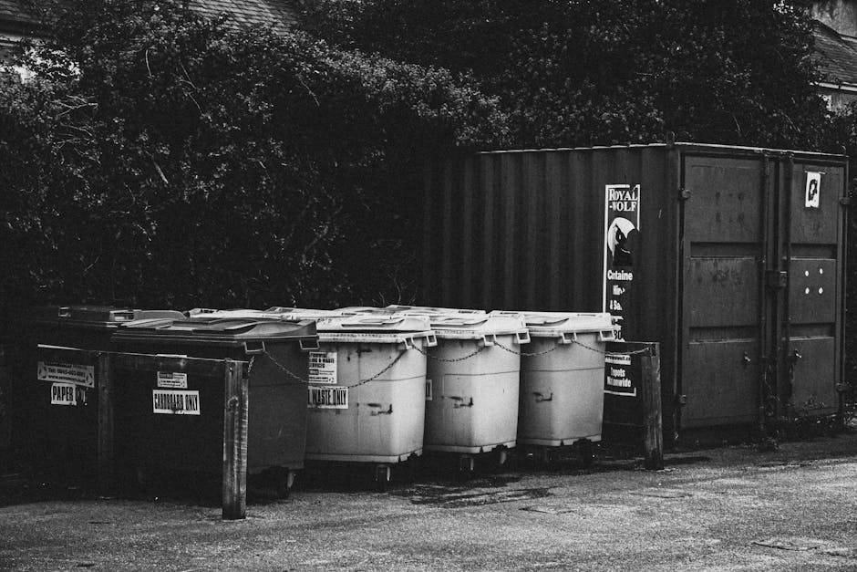 A large, green industrial waste disposal bin filled with various household and packaging materials, including cardboard boxes, plastic waste, and some metal items. The bin is placed outdoors on a paved area, with a mixture of opened and crushed cardboard boxes spilling over the top. In the background, there are additional cardboard boxes, some wooden pallets, and a street scene with trees and buildings, indicating a busy urban environment. The image captures a home relocation or moving process involving loading or disposal of bulky waste during a house removal or packing session, as part of the services provided by Man with Van Kingsbury.