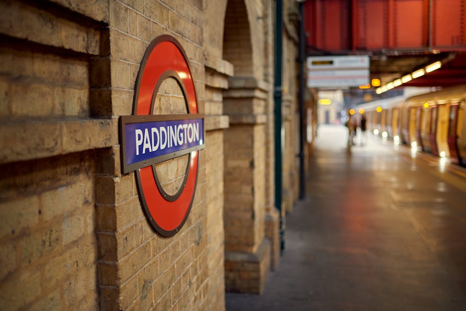 Inside the underground station at Kingsbury, the image shows a platform with a few passengers walking along the yellow tactile paving next to the train tracks. The platform is covered by a curved roof structure made of transparent panels supported by metal beams, allowing natural light to illuminate the area. On the left side, advertisements are visible on the wall, and a sign indicating the station's name and nearby bus routes is seen above the platform. To the right, a staircase leads up to the station entrance, with a prominent yellow 'Way out' sign and directional arrow pointing upwards. The station features tiled walls with illuminated signage, and the train tracks extend into a dark tunnel at the far end of the platform. This setting reflects the typical environment where local house removals and furniture transport are coordinated, with easy access for loading and unloading large items using dedicated transport services like those provided by Man with Van Kingsbury, especially during home relocation or packing and moving processes.