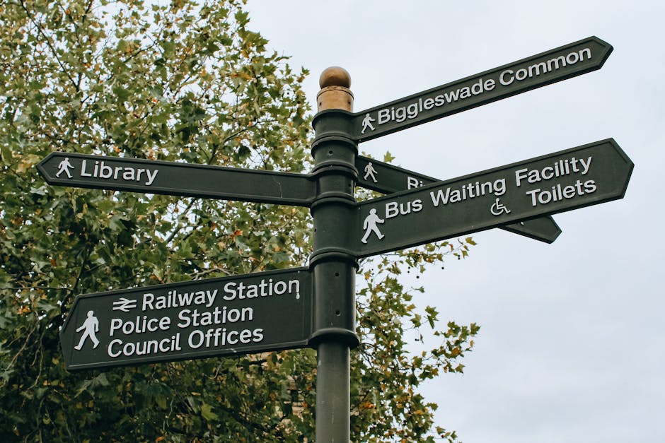 A black multi-directional street signpost standing outdoors during daytime, with six rectangular signs attached to a central pole. The signs point in various directions and display white text and symbols indicating the locations of Biggleswade Common, a library, a bus waiting facility and toilets, a railway station, a police station, and council offices. Some signs include pedestrian icons, while others feature wheelchair accessibility symbols. The background includes a tree with green leaves and an overcast sky, and the area appears to be a public space or walkway. The image reflects typical signage seen near urban or town centers, relevant for home relocation and transportation planning, as seen in the context of house removals and moving services provided by Man with Van Kingsbury.