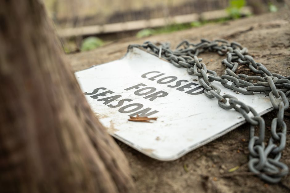 A weathered white metal sign reading 'Closed for Shason' lies on the ground outdoors, partially obscured by a thick metal chain laid across it. The sign appears dirty and slightly bent, with evidence of rust and dirt accumulation, and a small brown leaf resting on its surface. The scene is set in a natural environment, with the ground composed of soil and small pebbles, and a wooden fence or structure visible in the background. To the left, there is a blurred tree trunk, suggesting the sign is placed near a tree or in a garden area. The lighting is natural, indicating daytime. The chain looks sturdy and is possibly used for securing or blocking access, consistent with typical house or property management practices. This image reflects aspects of property security and access control, relevant to furniture transport, loading processes, or site management in house removals, such as those managed by Man with Van Kingsbury.