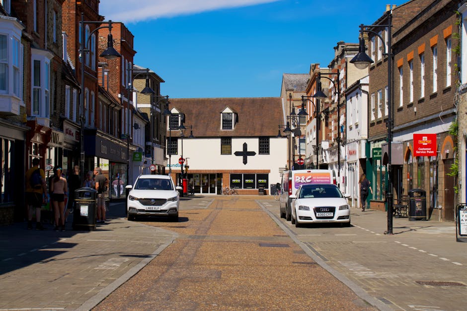 A wide view of a busy pedestrianised street in Kingsbury, showing shops on both sides, including a retail store and a customer service point, with several parked cars and vans on the paved street. Visible are groups of people, some standing near shop entrances and others walking along the pavement. The street is lined with black street lamps with downward-facing lights, and there are trash bins positioned along the curbs. In the background, a building with a distinct white façade and a black cross symbol on the front is visible, likely a healthcare or pharmacy outlet. The scene is illuminated by bright daylight, with a clear blue sky overhead. This setting could relate to home relocation or furniture transport activities conducted by Man with Van Kingsbury, involving loading or unloading items during a house move or delivery process within a busy commercial area.