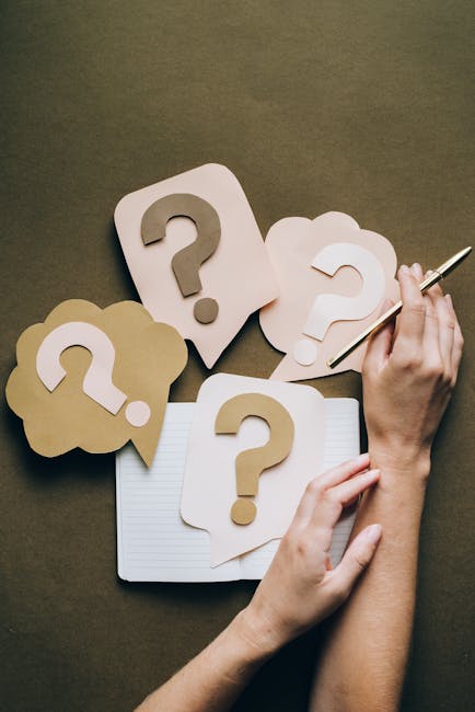 A person’s hands are visible, one hand holding a pen and the other supporting a notebook. The workspace features several paper cutouts shaped like speech bubbles and cloud shapes, each with a large question mark printed on them, arranged on a brown surface. Some of these paper questions are layered, with one or two placed on top of the notebook, which is open and blank. The scene appears to be set in an indoor environment with soft lighting, possibly relating to planning or decision-making for a house removal, such as quotes or pricing inquiries, connected to the services offered by Man with Van Kingsbury. The image emphasizes the theme of questions and clarification, relevant to understanding removals pricing and services in Kingsbury.