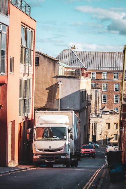 A white Renault cargo van is parked on a narrow urban street beside a multi-storey residential building with orange and beige exterior walls, large windows, and a balcony. The van is positioned close to the kerb, partially blocking the roadway, and appears to be preparing for a home relocation. Surrounding the van, there are several cardboard boxes and wrapped furniture, indicating packing and moving activities. A red car is parked further down the street, and bins are visible on the pavement nearby. The scene is illuminated by daylight, with clear blue skies above, and the adjacent buildings display a mixture of modern and older architectural styles. This setting exemplifies the logistical process of furniture transport and loading within a city environment, consistent with services offered by Man with Van Kingsbury for house removals and moving logistics.