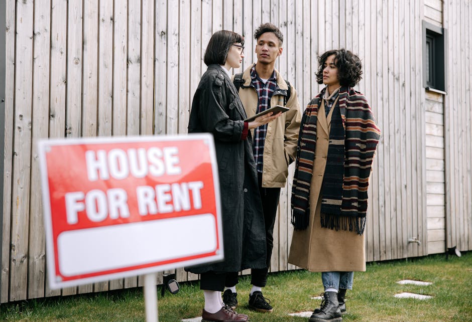 Three individuals standing outside a wooden house with vertical paneling, engaged in a discussion about a house rental, with two of them holding a clipboard, likely related to house removals or property management. The scene is set on a grassy area with some stepping stones and a small window on the house visible in the background. A prominent red and white 'House For Rent' sign is positioned in the foreground, partially out of focus, indicating a property available for lease. The individuals are dressed in casual clothing suitable for moving or property visits, and the scene is lit with natural daylight. The context suggests a home relocation or property viewing scenario, aligning with services offered by Man with Van Kingsbury for removals and moving logistics related to eviction or urgent housing situations in NW9, Kingsbury.