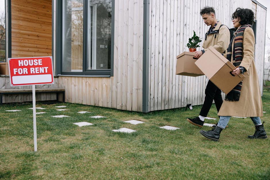 Two individuals dressed in coats are walking across a lawn next to a modern wooden house, carrying cardboard boxes filled with household items, including a potted plant. They are approaching a van for a home relocation or furniture transport service, with the side door or loading area visible in the background. The scene is outdoors in daytime, with a red and white 'House For Rent' sign planted in the grass near the house. The ground features neatly arranged paving stones leading to the house entrance, and the area is well-lit with natural light. The presence of packing materials and the action of loading or moving items suggest a professional removals process, supported by companies like Man with Van Kingsbury for tenants facing eviction or urgent moves, especially relevant for NW9 residents.