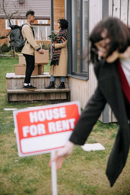 Three individuals standing outside a wooden house with vertical paneling, engaged in a discussion about a house rental, with two of them holding a clipboard, likely related to house removals or property management. The scene is set on a grassy area with some stepping stones and a small window on the house visible in the background. A prominent red and white 'House For Rent' sign is positioned in the foreground, partially out of focus, indicating a property available for lease. The individuals are dressed in casual clothing suitable for moving or property visits, and the scene is lit with natural daylight. The context suggests a home relocation or property viewing scenario, aligning with services offered by Man with Van Kingsbury for removals and moving logistics related to eviction or urgent housing situations in NW9, Kingsbury.