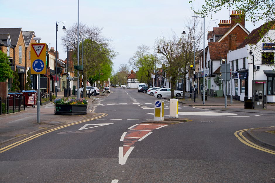 Image of a narrow street in Kingsbury lined with three- to four-storey brick houses featuring white-framed windows and chimneys, taken during daylight with a partly cloudy sky. A black vehicle is park