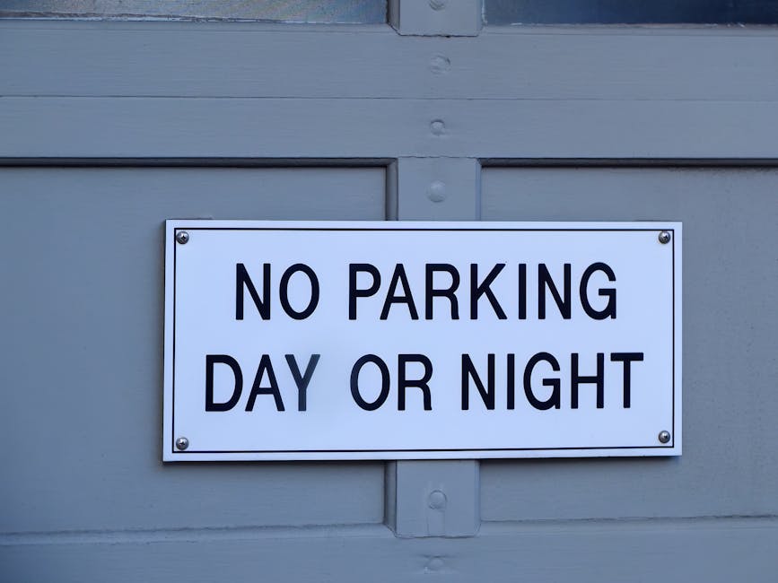 A rectangular white metal sign with bold black lettering reading 'NO PARKING DAY OR NIGHT' is mounted on a grey wooden door or garage door. The sign is attached with four screws, one at each corner. The door features horizontal and vertical panel details, and the surface has a smooth finish. The environment appears to be an outdoor or semi-outdoor setting, with natural lighting highlighting the sign and door surface. The sign's placement indicates a restriction on vehicle parking on the driveway or property frontage, which is relevant for residents and professional removal services like Man with Van Kingsbury when planning home relocation or furniture transport, especially during loading and unloading phases.