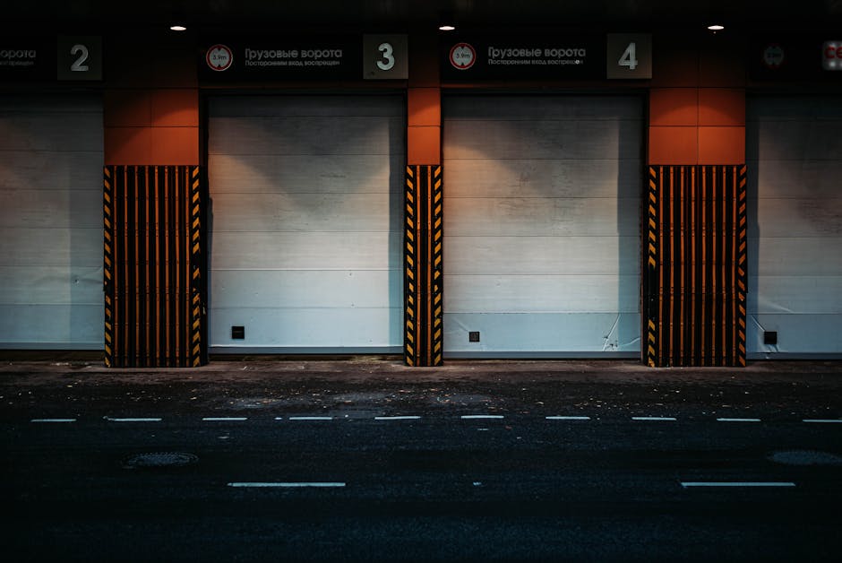 An empty loading bay area in a building with three large white garage doors, numbered 2, 3, and 4, each framed by black and yellow hazard striped protective barriers. The scene appears to be part of a commercial or industrial property, with a dark asphalt road in the foreground that has white dashed lines marking parking or loading zones. The building’s exterior wall is light-colored, and there are signs above each loading door indicating their numbers, including some text in Russian. The lighting is subdued, suggesting early morning or late evening, and the space is clear of vehicles or equipment, prepared for a home relocation or furniture transport operation by professionals like Man with Van Kingsbury, supporting efficient packing and moving processes within tightly controlled parking and loading rules.