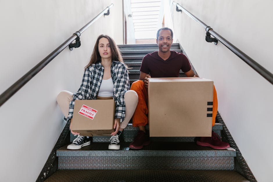 A young woman and a man are seated on a staircase inside a property, each holding packed cardboard boxes for a home relocation. The woman, wearing a black-and-white plaid shirt, white top, and sneakers, holds a smaller cardboard box with a red 'fragile' sticker, while the man, dressed in a maroon T-shirt and orange pants, holds a larger plain cardboard box. The staircase has a dark, textured rubber step surface, white walls, and metal handrails on both sides. Natural light from a nearby window illuminates the area, highlighting the packing and moving process taking place. This scene, captured during the loading process, reflects the careful handling of belongings by professionals such as Man with Van Kingsbury, ensuring furniture transport and packing are efficiently managed during house removals on Kingsbury terraces.